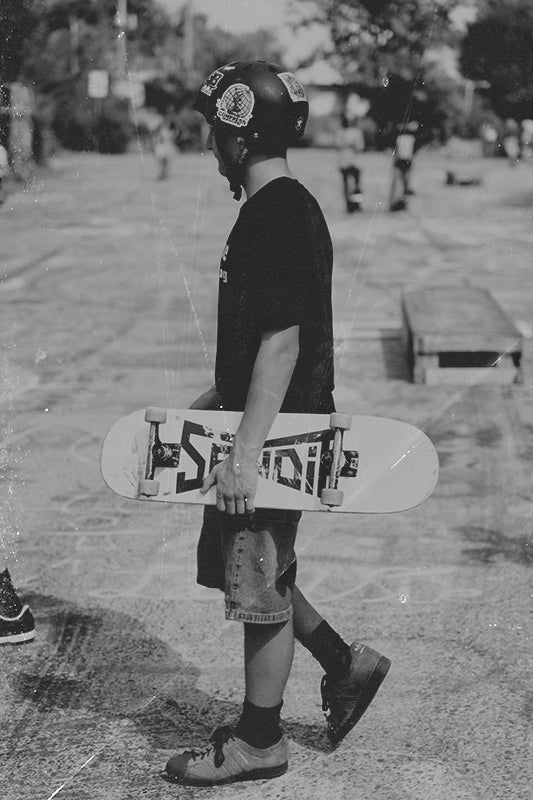 A black and white photograph of a skateboarder walking outdoors while holding a skateboard with graphic text on the underside. The person is wearing a helmet, dark t-shirt, shorts, socks, and skate shoes in a skatepark setting.