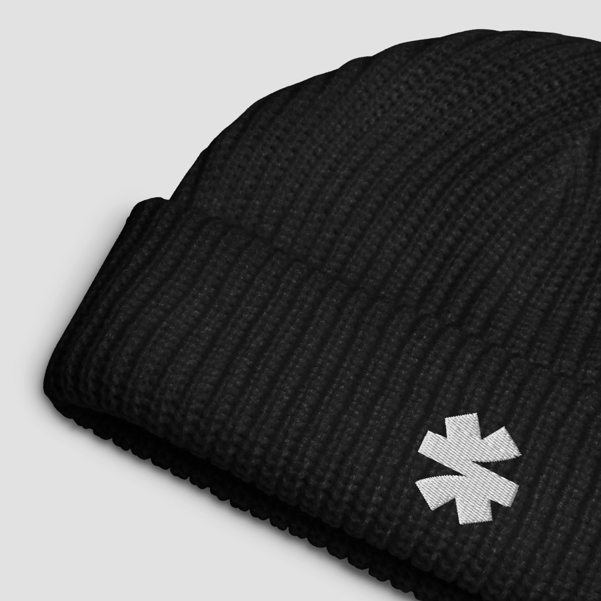 A close-up of a black ribbed knit beanie hat featuring a small white embroidered asterisk logo on the cuff, set against a light gray background.
