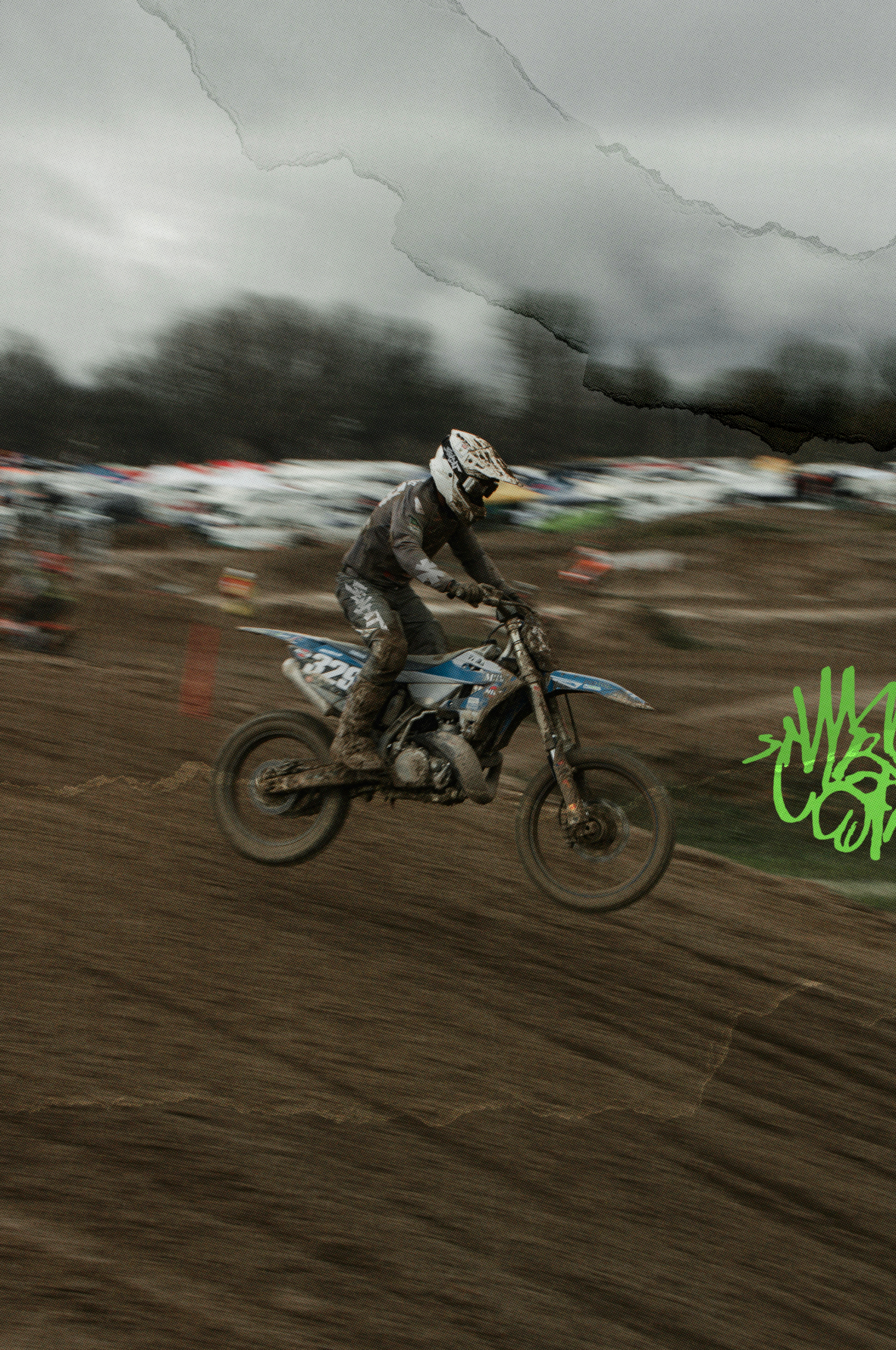 A motocross rider in full protective gear jumps a blue and white dirt bike over a muddy track under an overcast sky, with blurred trees and parked vehicles in the background.