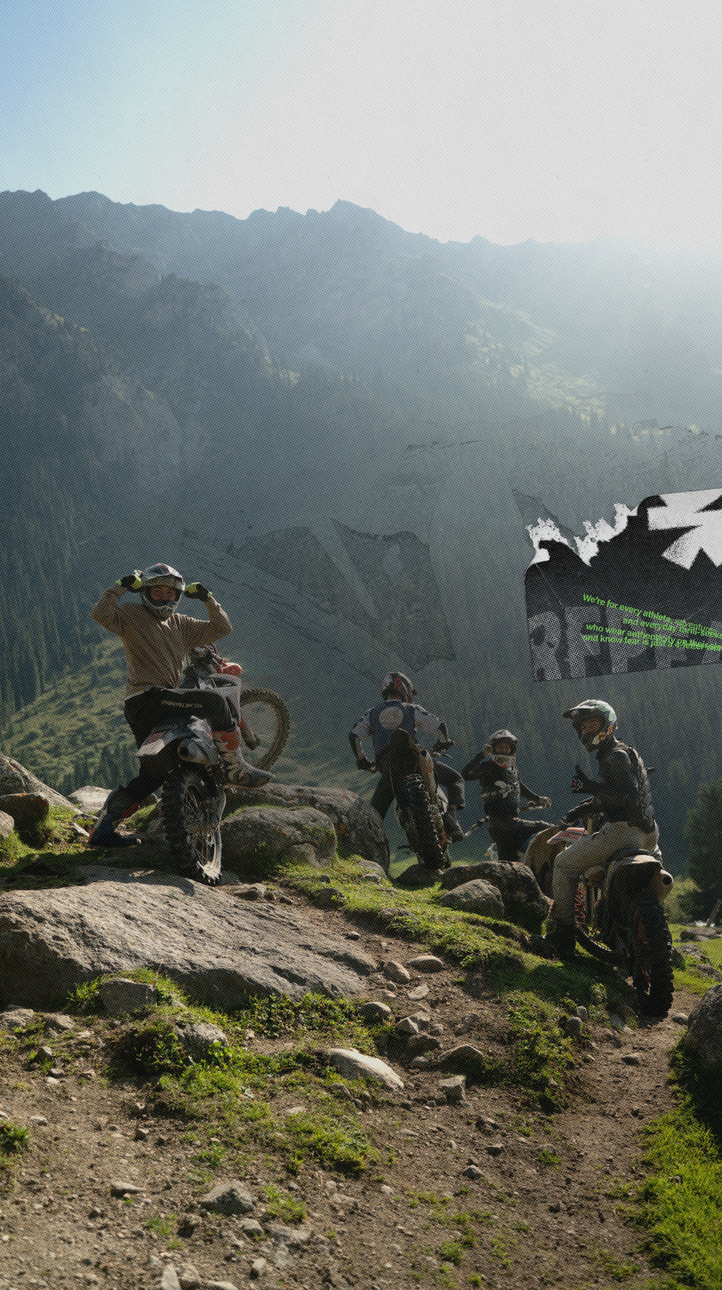 Several riders on off-road motorcycles navigate a rocky mountain trail with steep, rugged peaks in the background under a hazy sky.