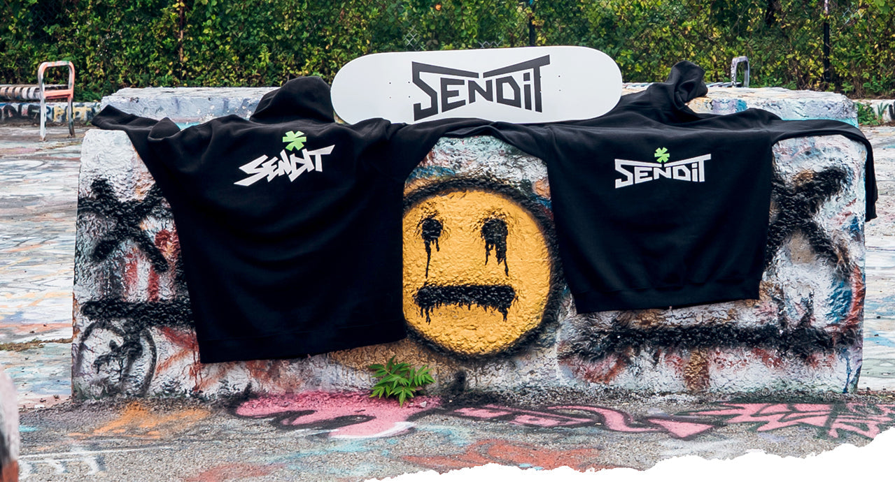 two black sweatshirts and a white skateboard deck with Send It logos on them laid out on a concred block outdoors with graffiti painted on all surfaces