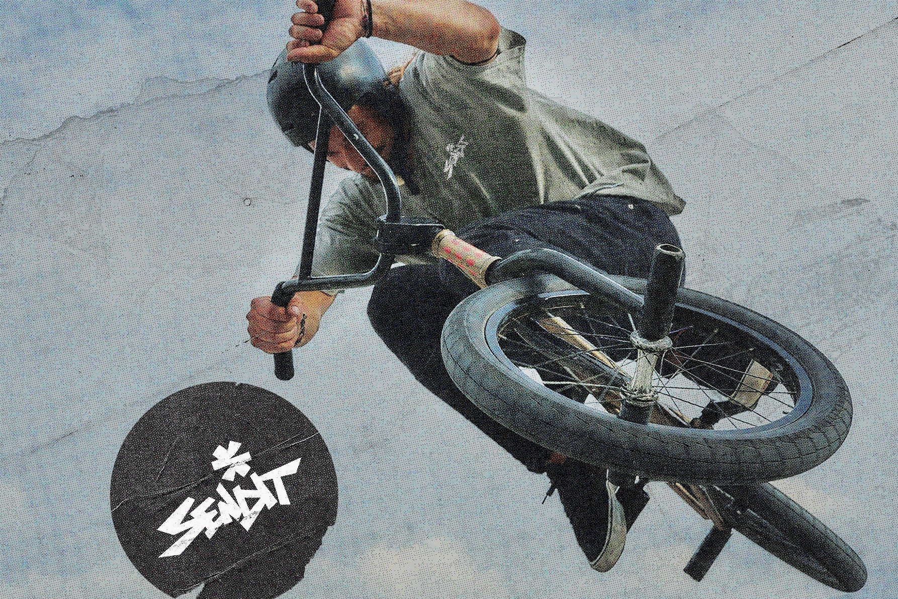 A BMX rider in mid-air performing a trick on a black BMX bike against a concrete skatepark background. The rider is wearing a gray t-shirt, black helmet, and black pants.